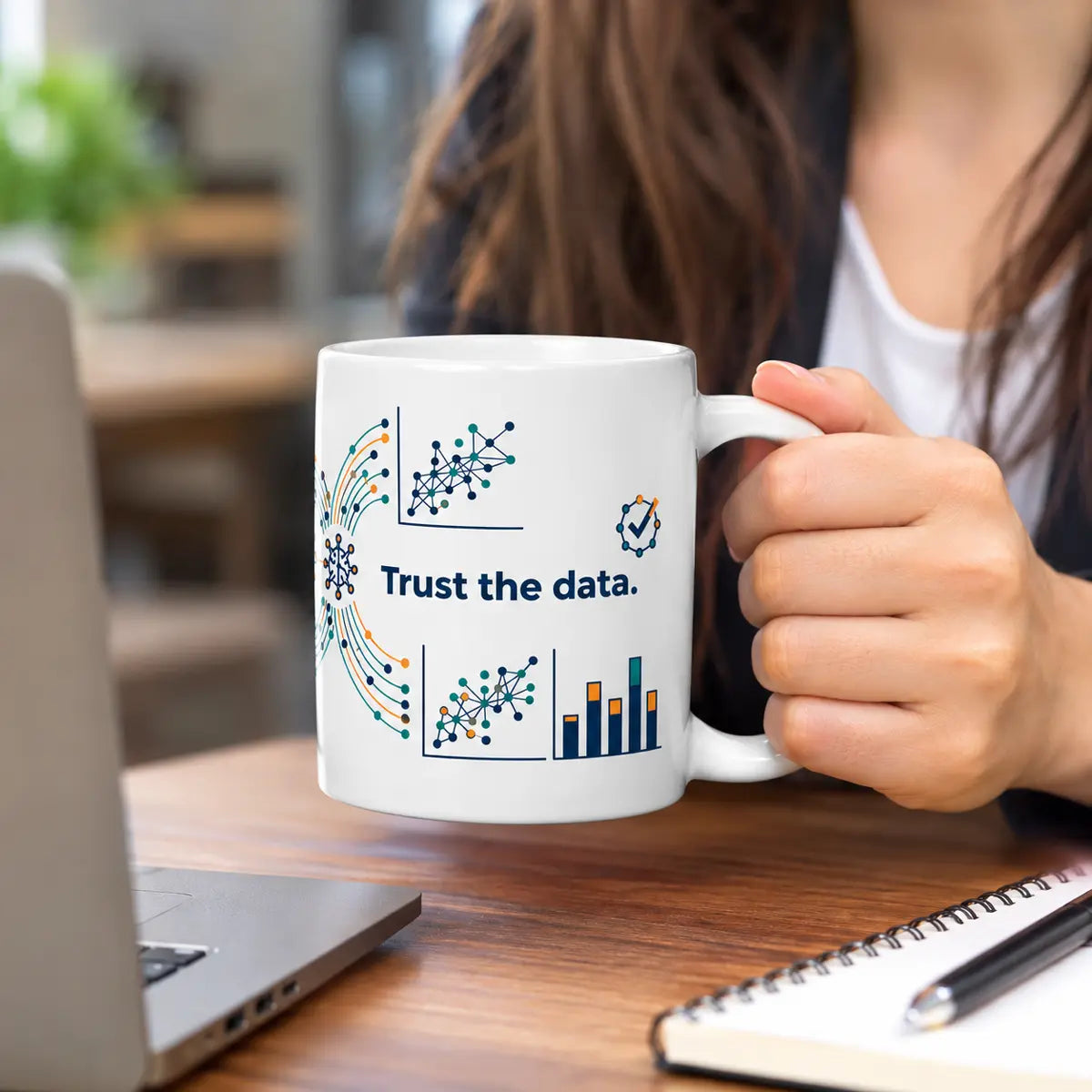 Woman holding a white ceramic coffee mug printed with data science charts, neural network graphs, and the phrase Trust the data at a desk.