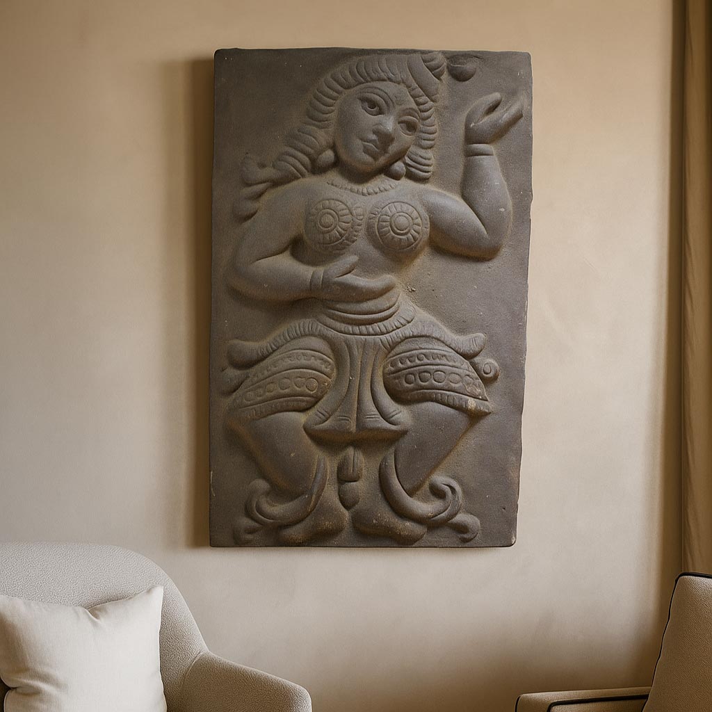 A lifestyle photograph showcasing the handcrafted terracotta dancer relief panel mounted on a neutral beige wall. The artwork is displayed above a contemporary armchair, illustrating how this traditional piece of Indian temple art adds rustic heritage and cultural depth to a modern living space.