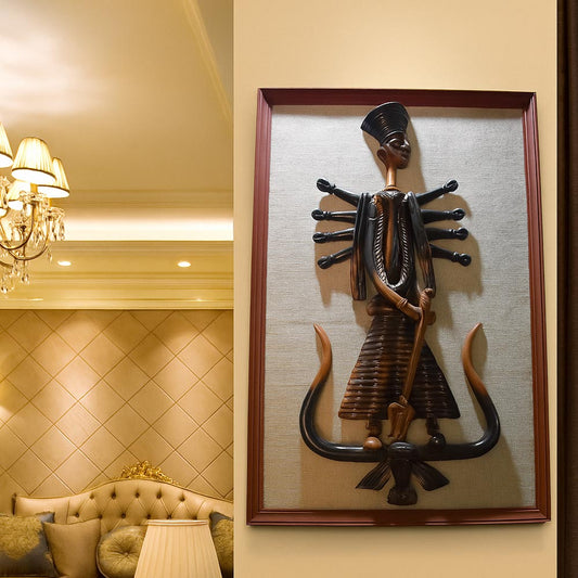 A lifestyle photograph displaying a framed handcrafted terracotta wall hanging of Goddess Durga. The traditional ten-armed clay figure is mounted on a light wall in a luxurious room setting featuring a crystal chandelier and tufted furniture, illustrating how this ethnic art piece enhances sophisticated home interiors.