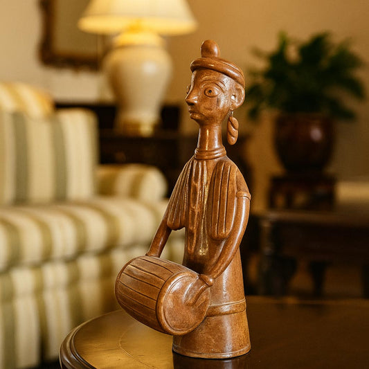 A close-up side view of a terracotta musician figurine placed on a wooden coffee table in a cozy living room setting. The background is softly blurred, showing a sofa and lamp, highlighting the figurine as a sophisticated home accent.