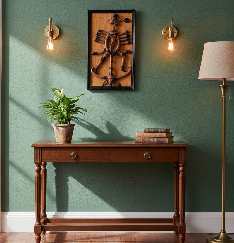 An interior design shot featuring the handcrafted Bankura style terracotta Maa Durga wall hanging mounted on a textured sage green wall. The artwork is illuminated by vintage brass wall sconces and positioned above a dark wooden console table styled with a potted plant and a stack of antique books. The setting showcases a blend of traditional Indian heritage and sophistication in modern home styling.