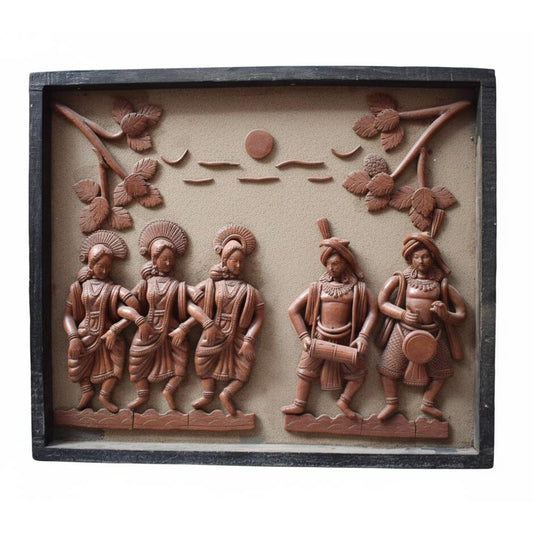 A horizontal handcrafted terracotta wall hanging depicting a lively tribal scene. The relief sculpture features three women in traditional attire dancing in rhythm on the left, accompanied by two men playing percussion drums on the right. The scene is set against a textured beige background with stylized tree branches and a sun motif, enclosed in a dark wooden frame.