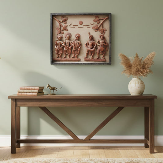 A serene lifestyle photograph featuring the handcrafted terracotta tribal wall art hanging on a sage green feature wall. The artwork is styled above a dark wooden console table with cross-legs, accompanied by a stack of vintage books, a small brass horse figurine, and a beige vase filled with dried pampas grass.