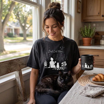 Woman wearing a washed black Best Cat Mom crop top while holding a matching coffee mug with a black cat on her lap in a bright kitchen lifestyle setting.