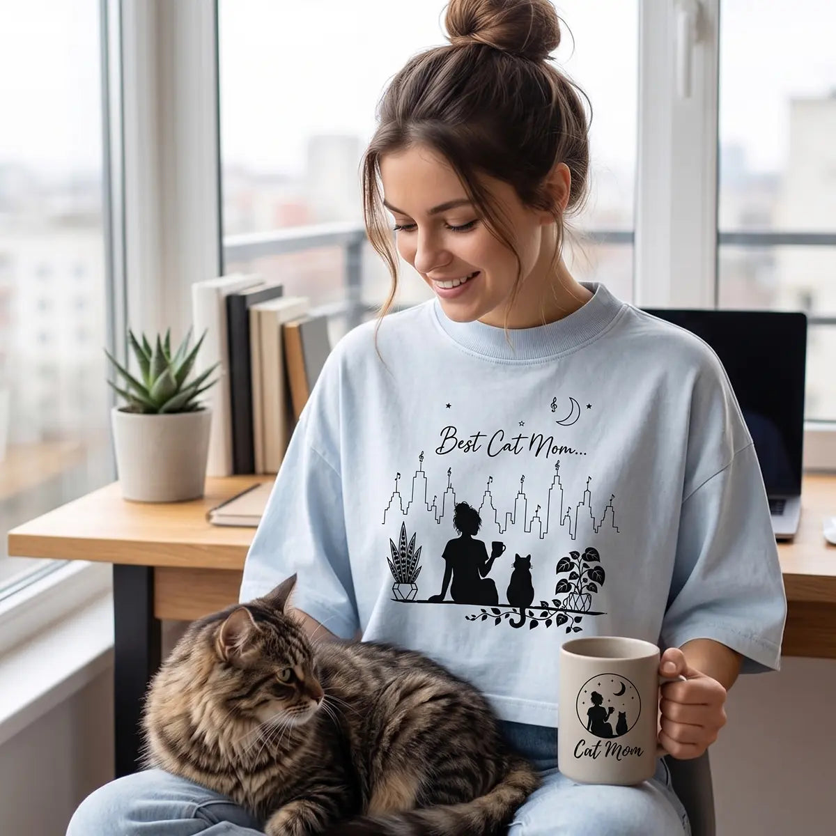 Woman wearing a light washed denim Best Cat Mom crop top while holding a coffee mug with a tabby cat on her lap in a bright home office.