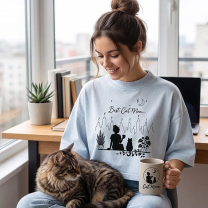 Woman wearing a light washed denim Best Cat Mom crop top while holding a coffee mug with a tabby cat on her lap in a bright home office.