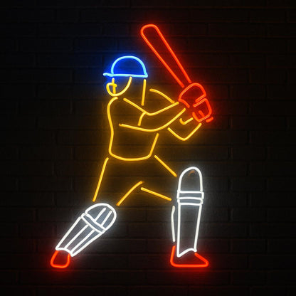 A colorful LED neon sign featuring a cricket batsman in action. The design showcases distinct colors with a blue helmet, red bat and gloves, golden-yellow body, and white leg pads, all illuminated against a dark brick wall background. Ideal for cricket fans and sports-themed interiors.