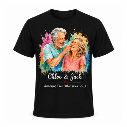 A black unisex t-shirt featuring a custom watercolor portrait of a smiling older couple surrounded by vibrant paint splatters, with funny anniversary text below. Free shipping by Incotta.