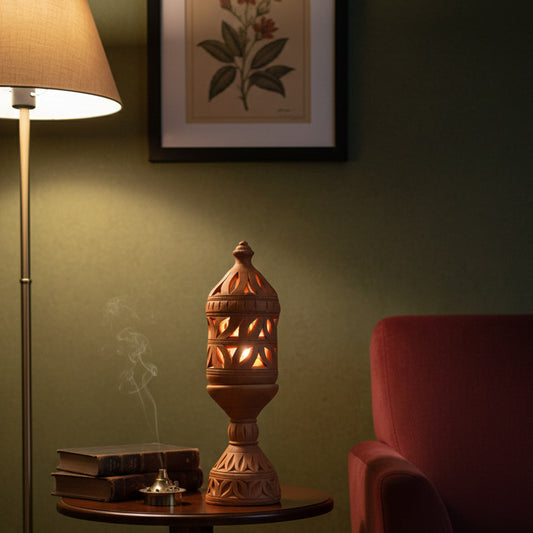 A bright lifestyle photograph of a terracotta lantern centerpiece on a wooden table. The setting includes a potted fern, a stack of vintage books, a brass incense burner with smoke, and a table lamp with a floral shade against a sage green wall.