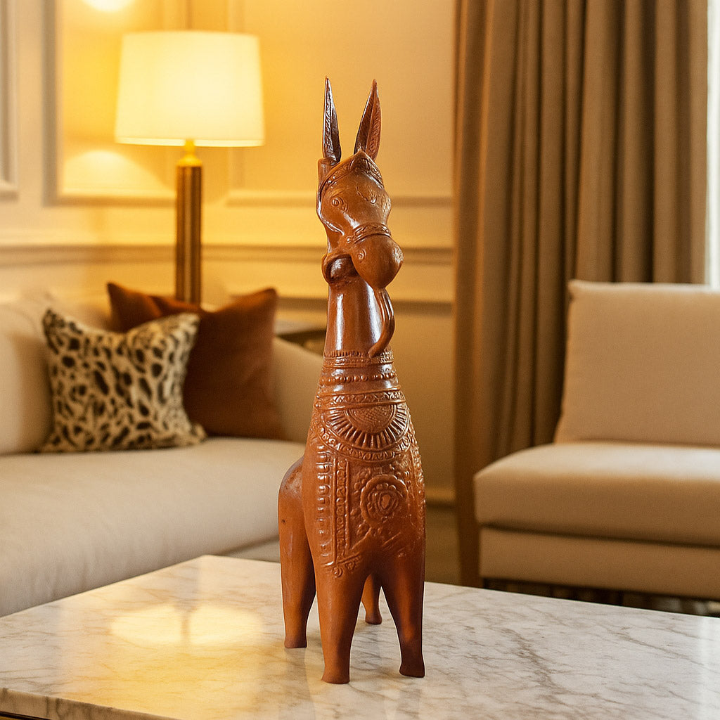 A lifestyle photograph of a traditional Bankura terracotta horse statue displayed on a marble coffee table. The handcrafted clay sculpture is highlighted by warm ambient lighting in a sophisticated living room setting, showcasing it as a premium piece of ethnic home styling.