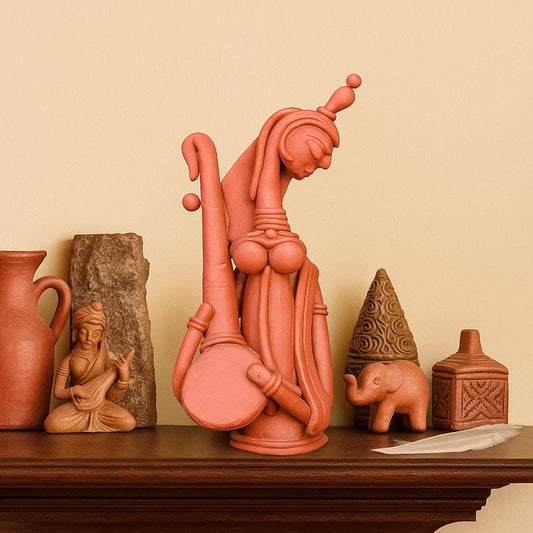 A serene lifestyle photograph featuring a handcrafted terracotta Saraswati statue playing the Veena, displayed on a dark wooden shelf. The goddess figurine is styled alongside complementary earthy decor elements—including clay pots, a miniature elephant, and stone accents—creating a culturally rich focal point against a warm beige background.