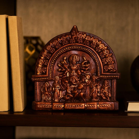 A lifestyle photograph showcasing the handcrafted terracotta Mahishasuramardini Durga idol with a dark antique finish. The sculpture is displayed on a wooden shelf alongside hardcover books, demonstrating how this traditional spiritual piece serves as a sophisticated accent for a home library or study.