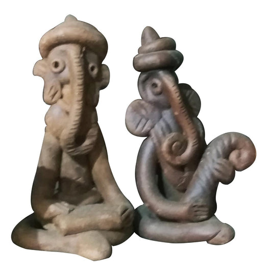 A studio shot featuring a pair of handcrafted terracotta figurines depicting stylized Lord Ganesha. The set includes two seated idols, one in a meditative posture and the other playing a curved musical instrument, showcasing traditional tribal design and earthen clay textures isolated against a white background.