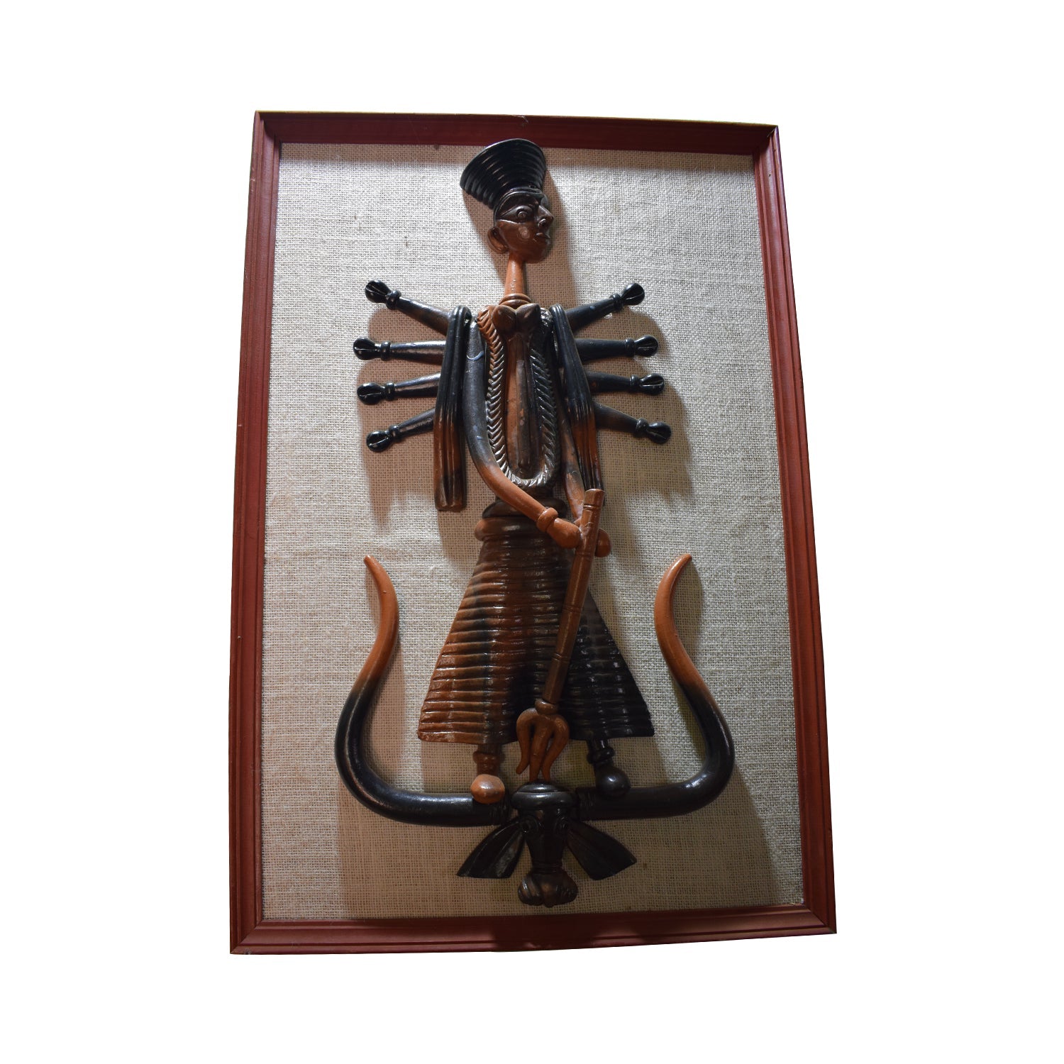 A front view of a handcrafted terracotta wall hanging depicting Goddess Durga, mounted on a textured beige fabric background within a wooden frame. This stylized clay sculpture features the ten-armed goddess standing on a buffalo demon motif, showcasing a unique blend of dark and reddish-brown earthenware tones.
