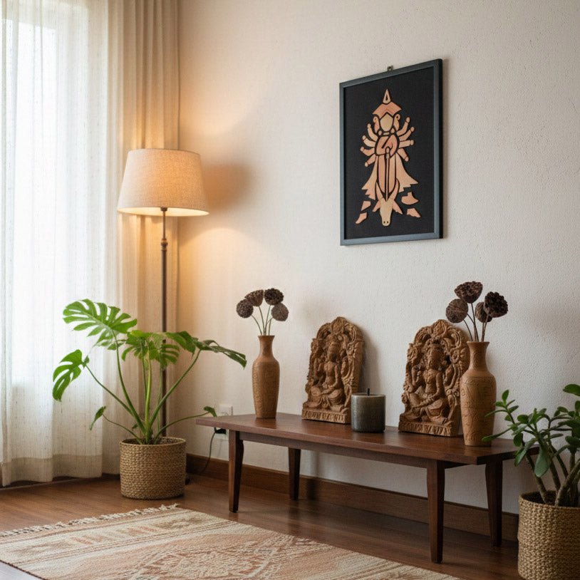 A serene interior setting featuring the modern geometric terracotta Goddess Durga wall hanging mounted on a white wall. The artwork is styled above a low wooden bench displaying carved ethnic deities, earthenware vases with dried botanicals, and a candle. A floor lamp and large Monstera plant in a woven basket add warmth and natural texture to this contemporary spiritual corner.