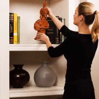 An animated view demonstrating how to style the handcrafted terracotta musician doll on a white living room bookshelf. The visual shows a person placing the tribal clay figurine next to books and vases, highlighting its perfect size and aesthetic appeal for modern shelf decor.