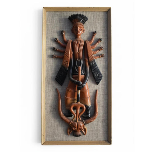 An exquisite handcrafted terracotta wall mural depicting a stylized, tribal interpretation of Goddess Durga as Mahishasura Mardini. The ten-armed deity, finished in dual-tone earthen red and black, stands triumphantly over the buffalo demon while holding a trident. The clay relief is mounted on a textured natural fabric backing and enclosed in a light wooden frame, making it a perfect statement piece for ethnic home decor.