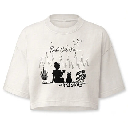 Stay cozy with this personalized minimalist best cat mom crop top in vintage white. Features a black line-art silhouette of a girl holding a coffee mug alongside a cat, potted plants, and a city skyline.