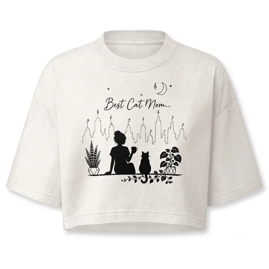 Stay cozy with this personalized minimalist best cat mom crop top in vintage white. Features a black line-art silhouette of a girl holding a coffee mug alongside a cat, potted plants, and a city skyline.