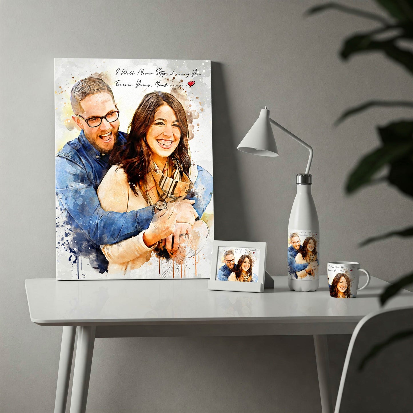 A display of custom photo merchandise on a white desk, featuring a watercolor-style portrait of a happy couple. The collection includes a large vertical canvas print with the romantic text "I Will Never Stop Loving You," alongside a matching printed water bottle, coffee mug, and a small framed photo, making it an ideal anniversary or Valentine's Day gift set.