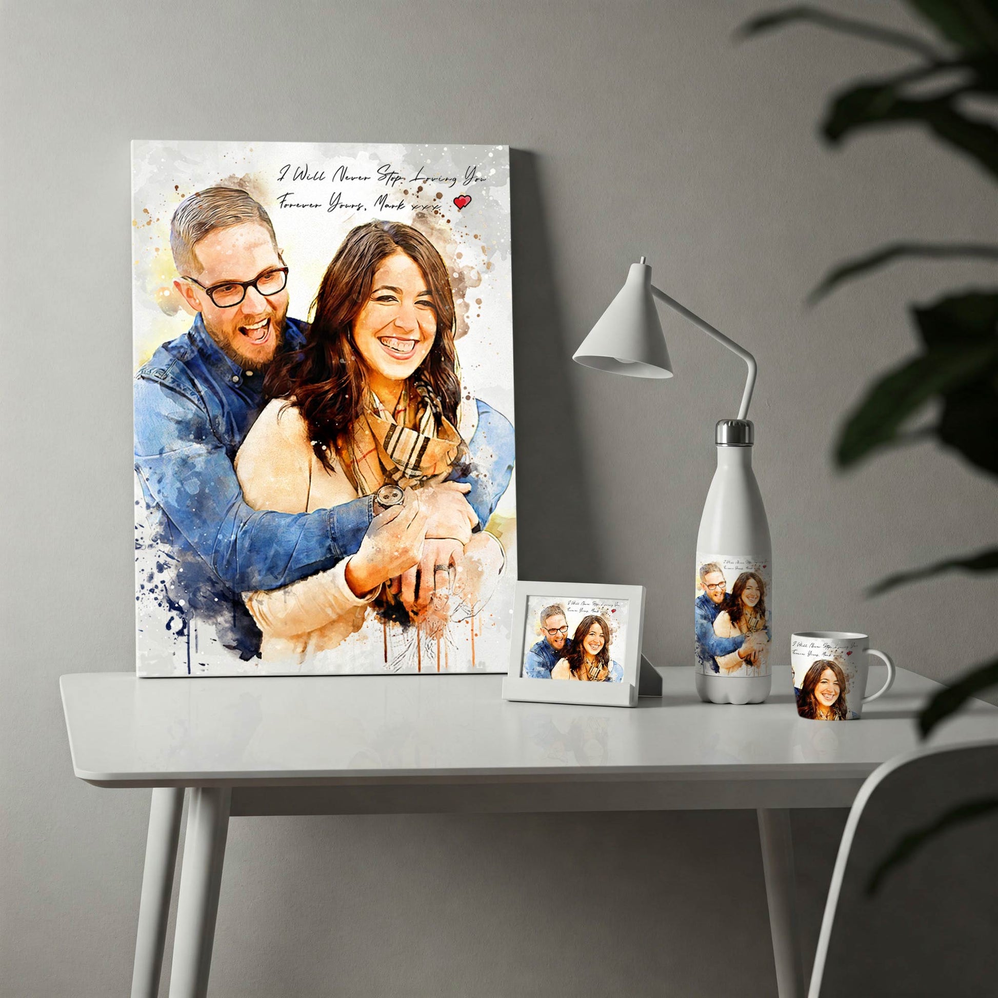 A display of custom photo merchandise on a white desk, featuring a watercolor-style portrait of a happy couple. The collection includes a large vertical canvas print with the romantic text "I Will Never Stop Loving You," alongside a matching printed water bottle, coffee mug, and a small framed photo, making it an ideal anniversary or Valentine's Day gift set.