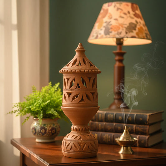 A cozy lifestyle photograph featuring a decorative terracotta lantern with cutout patterns standing on a polished wooden table. The styling includes a stack of vintage-style books, a brass incense burner with rising smoke, a small green potted plant, and a warm table lamp in the background, showcasing the lantern as a rustic home decor centerpiece.