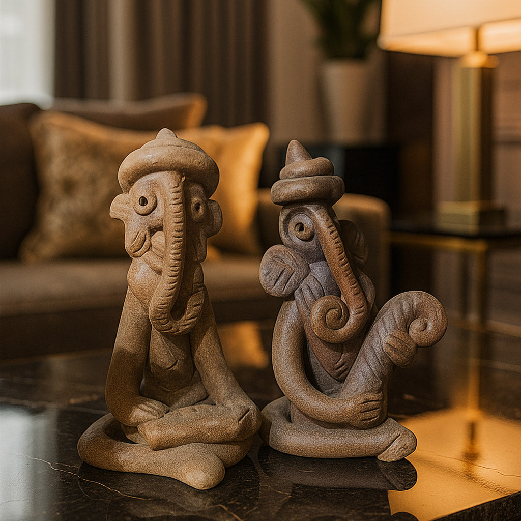 A high-quality lifestyle photograph displaying a pair of handcrafted terracotta Ganesha figurines placed on a reflective dark table surface. The image showcases the tribal-style idols, one meditating and one playing an instrument, in a warm, sophisticated living room setting with a beige sofa and lamp in the blurred background.