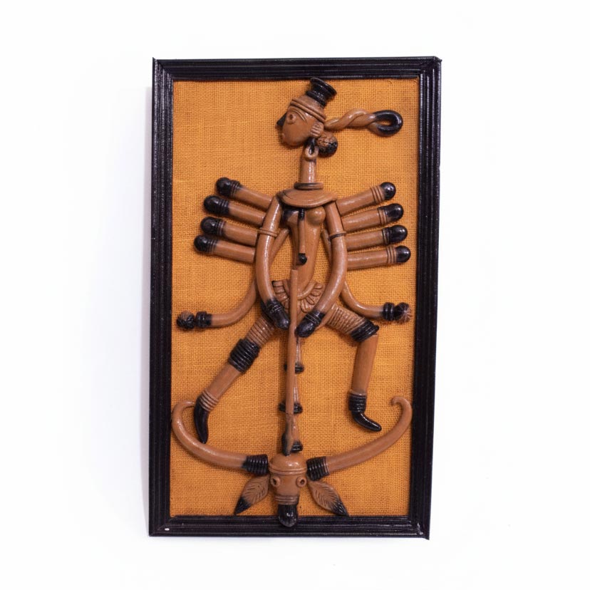 A front view of a framed terracotta wall hanging depicting Goddess Durga in the traditional Bankura folk art style of West Bengal. The stylized clay figure features elongated limbs and ten arms, portrayed in the Mahishasura Mardini pose (slaying the buffalo demon). The artwork uses natural brown clay tones with black painted accents on the hands, feet, and hair, mounted against a textured mustard-yellow fabric background within a black wooden frame.